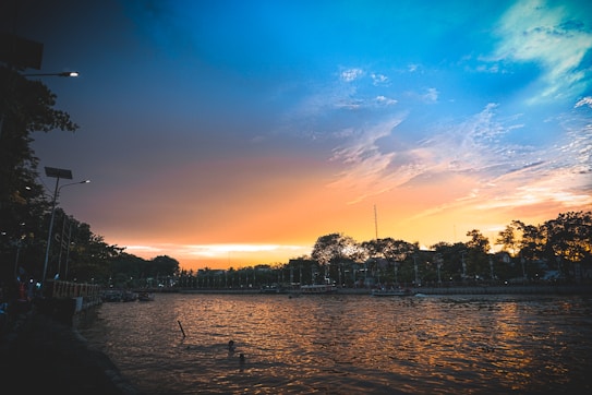 A scenic view of a river at sunset, with a vibrant sky transitioning from blue to warm orange hues. Trees line the horizon, silhouetted against the colorful sky, and a few streetlights are visible along the riverbank. The tranquil water reflects the sky's colors, enhancing the serene atmosphere.