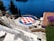 A tiled emblem with red and white checks and the words 'Hajduk Split' is prominently displayed on the ground of an open area overlooking a body of water. The scene includes nearby trees, a tiled roof of a building, and an array of bougainvillaea flowers. Stairs and railings descend towards the water, offering a view of the deep blue sea and a distant ship.