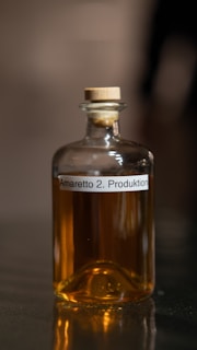 A small glass bottle with a wooden stopper contains a brown liquid. The label on the bottle reads 'Amaretto 2. Produktion'. The bottle is placed on a reflective surface, and the image is softly focused, emphasizing the warm hue of the liquid.