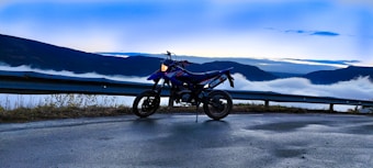 A motorcycle is parked on a road with a picturesque mountain backdrop. The sky is a soft blend of blue with clouds hovering over valleys below, creating a serene atmosphere. The motorcycle stands on an empty road bordered by a metal guardrail, surrounded by natural scenery.