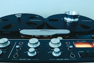 A vintage reel-to-reel tape recorder is displayed, featuring large black reels and metallic control knobs. The device includes a range of control dials and switches, conveying a sense of classic audio equipment.