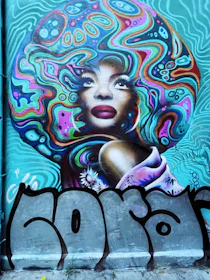 A vibrant mural featuring a stylized portrait of a person with bright, swirling patterns that resemble a large, colorful afro. The background and patterns are filled with psychedelic designs in blues, pinks, greens, and purples. The person has bold facial features, including full lips and prominent eyes, and there are floral elements and graffiti tags surrounding the image.