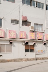 A building facade with multiple large pink signs displaying Japanese characters. Several windows are visible, some with reflective glass, and there is an air conditioning unit mounted near the top. Below the signs, there is a notice board with colorful illustrations and text.