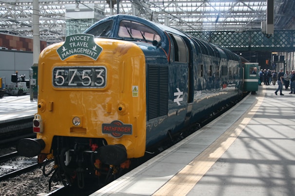 A vintage diesel locomotive with a yellow front and a dark blue body is stationed at a platform in a train station. The locomotive displays the numbers 5253 and has signs for 'Avocet Travel Management' and 'Pathfinder' on its front. The station platform is covered by a large glass and metal roof, and there are people standing in the background near the train. The environment suggests a classic, well-maintained transportation setting.