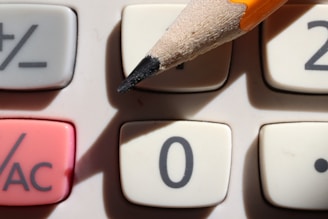 A close-up view of a calculator's keypad with a pencil poised on the number zero button. The keypad includes buttons for addition, subtraction, and an AC (all clear) function, which is highlighted in red.