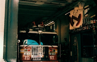 A fire truck is parked inside a dimly lit fire station garage. Above the vehicle, a large Ghostbusters logo featuring a cartoon ghost is prominently displayed on the wall. The garage is lined with bricks, and various posters and stickers decorate the area around the logo.