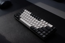 A mechanical keyboard with a minimalist design, featuring a combination of black and white keys. The keyboard is placed on a dark surface with soft lighting creating a shadow effect. The arrangement and style of the keys suggest a compact layout.