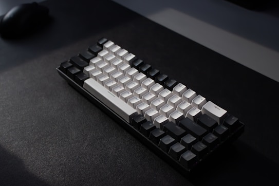 A mechanical keyboard with a minimalist design, featuring a combination of black and white keys. The keyboard is placed on a dark surface with soft lighting creating a shadow effect. The arrangement and style of the keys suggest a compact layout.