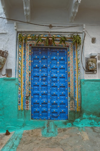 A vibrant blue door with intricate metal studs sits framed by a decorative, colorful border featuring floral and geometric designs. A garland of green leaves hangs above the door. The surrounding wall is painted in shades of teal and white, with some weathering and small embellishments.