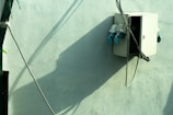 An electrical box mounted on a light green wall, casting a distinct shadow. Several cables are connected to the box, which appears partially open. The setting is outdoors, with sunlight creating sharp contrasts in the image.