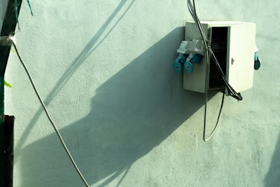 An electrical box mounted on a light green wall, casting a distinct shadow. Several cables are connected to the box, which appears partially open. The setting is outdoors, with sunlight creating sharp contrasts in the image.