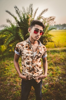 A young person stands confidently in a field with lush green grass and a palm tree in the background. They are wearing a patterned button-up shirt with rolled-up sleeves and sunglasses, projecting a casual and cool demeanor.