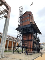 A large industrial structure made of red bricks and black metal beams, featuring a tower-like design with multiple windows and a metal staircase. The surrounding area is composed of cobblestone pathways and additional metal structures. A bird is flying above the building on a cloudy day.