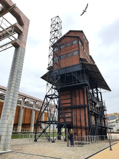 A large industrial structure made of red bricks and black metal beams, featuring a tower-like design with multiple windows and a metal staircase. The surrounding area is composed of cobblestone pathways and additional metal structures. A bird is flying above the building on a cloudy day.