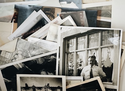 A collection of vintage black and white photographs is scattered across a surface. One prominent photograph shows a man in a shirt and tie standing in front of a building with decorative windows. Other photos include various figures and architectural elements.