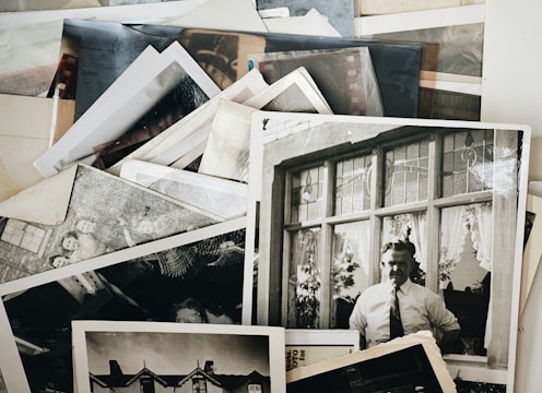 A collection of vintage black and white photographs is scattered across a surface. One prominent photograph shows a man in a shirt and tie standing in front of a building with decorative windows. Other photos include various figures and architectural elements.