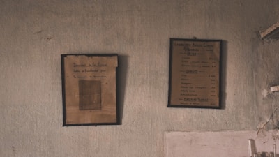 Two framed vintage documents are hanging on an aged, textured wall. The frames have a worn appearance, suggesting historical or antiquated content. The wall exhibits signs of wear and discoloration.