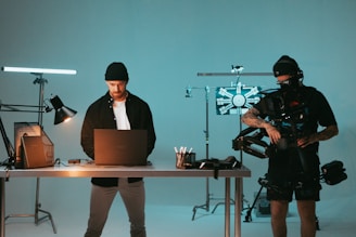 A man stands behind a desk with a laptop, wearing a beanie and casual clothing, while another individual operates a camera rig with professional filming equipment. The setting appears to be a studio with lighting equipment and various technical apparatus.