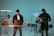 A man stands behind a desk with a laptop, wearing a beanie and casual clothing, while another individual operates a camera rig with professional filming equipment. The setting appears to be a studio with lighting equipment and various technical apparatus.