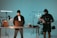 A man stands behind a desk with a laptop, wearing a beanie and casual clothing, while another individual operates a camera rig with professional filming equipment. The setting appears to be a studio with lighting equipment and various technical apparatus.