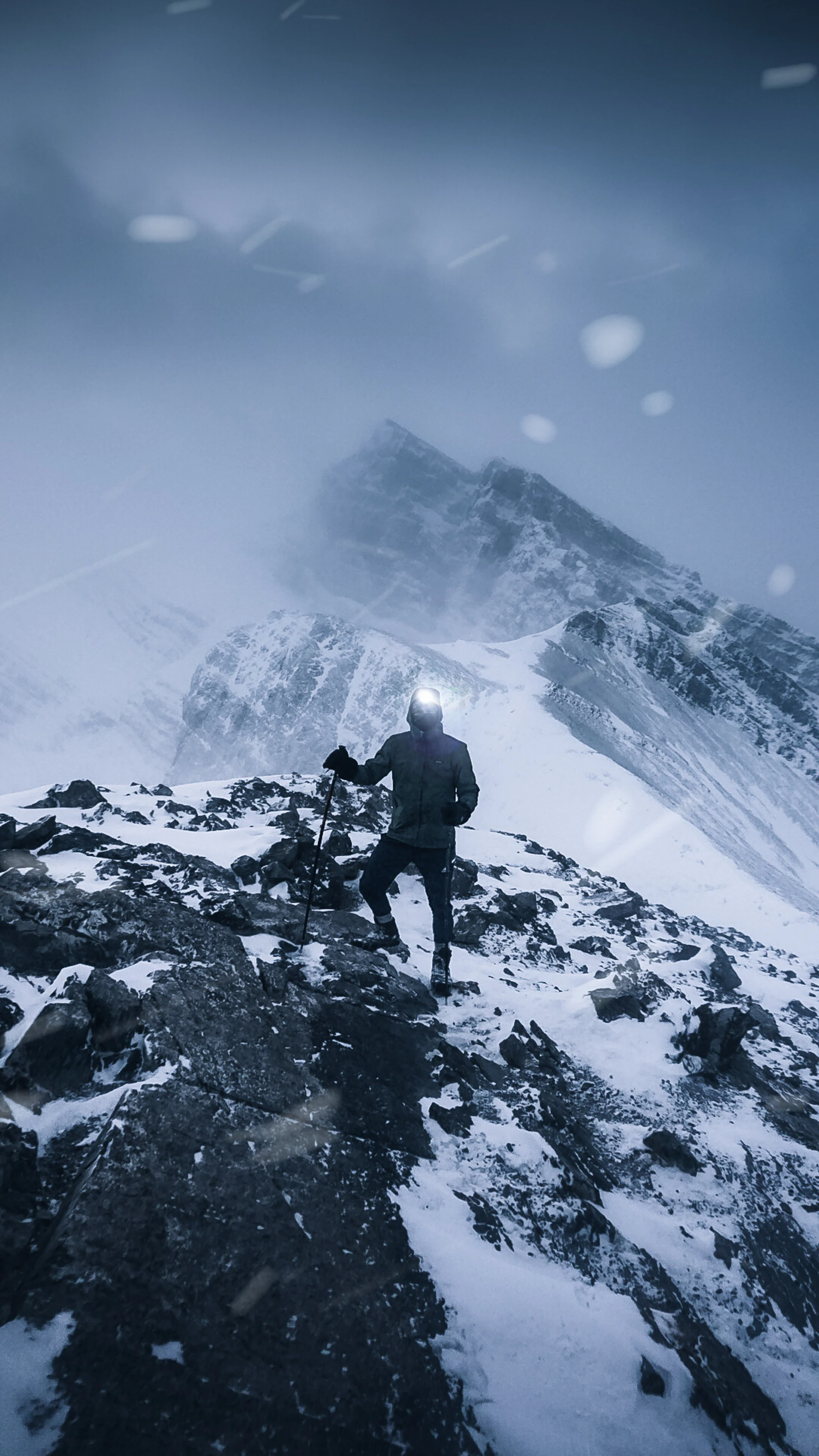 A figure with a tripod on a rocky snow ridge under a blowing summit
