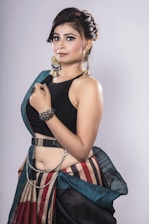 A woman with styled hair is wearing an elegant outfit featuring a black top and a colorful, striped skirt paired with a belt. She accessorizes with dramatic earrings, a bracelet, and a hand chain.
