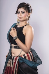 A woman with styled hair is wearing an elegant outfit featuring a black top and a colorful, striped skirt paired with a belt. She accessorizes with dramatic earrings, a bracelet, and a hand chain.