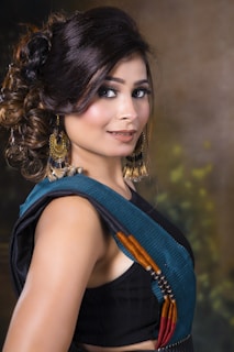 A woman with styled brown hair and elegant makeup, wearing large ornate earrings and a colorful sari. The background is a subtle blend of dark and warm tones, creating a sophisticated atmosphere.