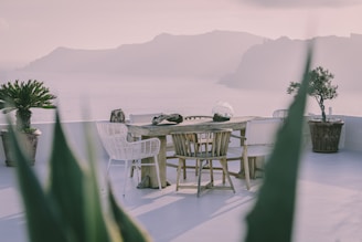 An elegant outdoor setting with a wooden table surrounded by wicker and wooden chairs on a patio overlooking a scenic ocean view with distant mountains. Potted plants adorn the area, and the atmosphere appears serene and calm.