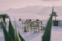 An elegant outdoor setting with a wooden table surrounded by wicker and wooden chairs on a patio overlooking a scenic ocean view with distant mountains. Potted plants adorn the area, and the atmosphere appears serene and calm.