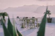 An elegant outdoor setting with a wooden table surrounded by wicker and wooden chairs on a patio overlooking a scenic ocean view with distant mountains. Potted plants adorn the area, and the atmosphere appears serene and calm.