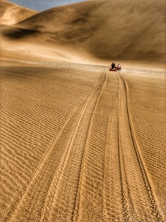 A vast expanse of sand dunes stretches into the distance, with two people riding red all-terrain vehicles along a narrow path. The sand appears smooth with visible tire tracks, suggesting recent activity.