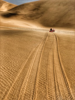A vast expanse of sand dunes stretches into the distance, with two people riding red all-terrain vehicles along a narrow path. The sand appears smooth with visible tire tracks, suggesting recent activity.