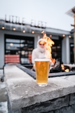 A glass of beer is placed on a stone surface in front of a fire pit with visible flames. In the background, there is a blurred figure wearing a white jacket and sunglasses, sitting on a bench. The building behind displays the words 'LITTLE FLEET' in illuminated letters.