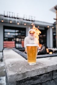 A glass of beer is placed on a stone surface in front of a fire pit with visible flames. In the background, there is a blurred figure wearing a white jacket and sunglasses, sitting on a bench. The building behind displays the words 'LITTLE FLEET' in illuminated letters.