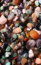 A collection of various polished gemstones in an array of colors and shapes. The stones are closely packed, showcasing shades of purple, orange, green, and black among others. The surface of each stone is smooth and shiny, reflecting light.