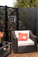A wicker patio chair with a light cushion and a red-orange floral pillow is positioned on a wooden deck. Next to the chair is a glass-topped side table. Behind the chair, there is a decorative panel with a leaf-like pattern cut out, and trees are visible in the background.