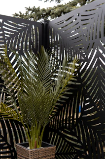 A tall plant with long green fronds is placed in a woven basket planter. The background features decorative metal panels with leaf patterns, creating an artistic and modern design. A hint of trees is visible in the upper background.