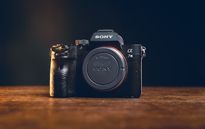 A black Sony camera with an E-mount and the model designation 'α7 III' is positioned on a wooden surface. The camera is central in the frame with a dark background, highlighting its details and prominent grip texture.