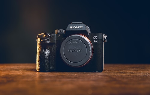 A black Sony camera with an E-mount and the model designation '&alpha;7 III' is positioned on a wooden surface. The camera is central in the frame with a dark background, highlighting its details and prominent grip texture.