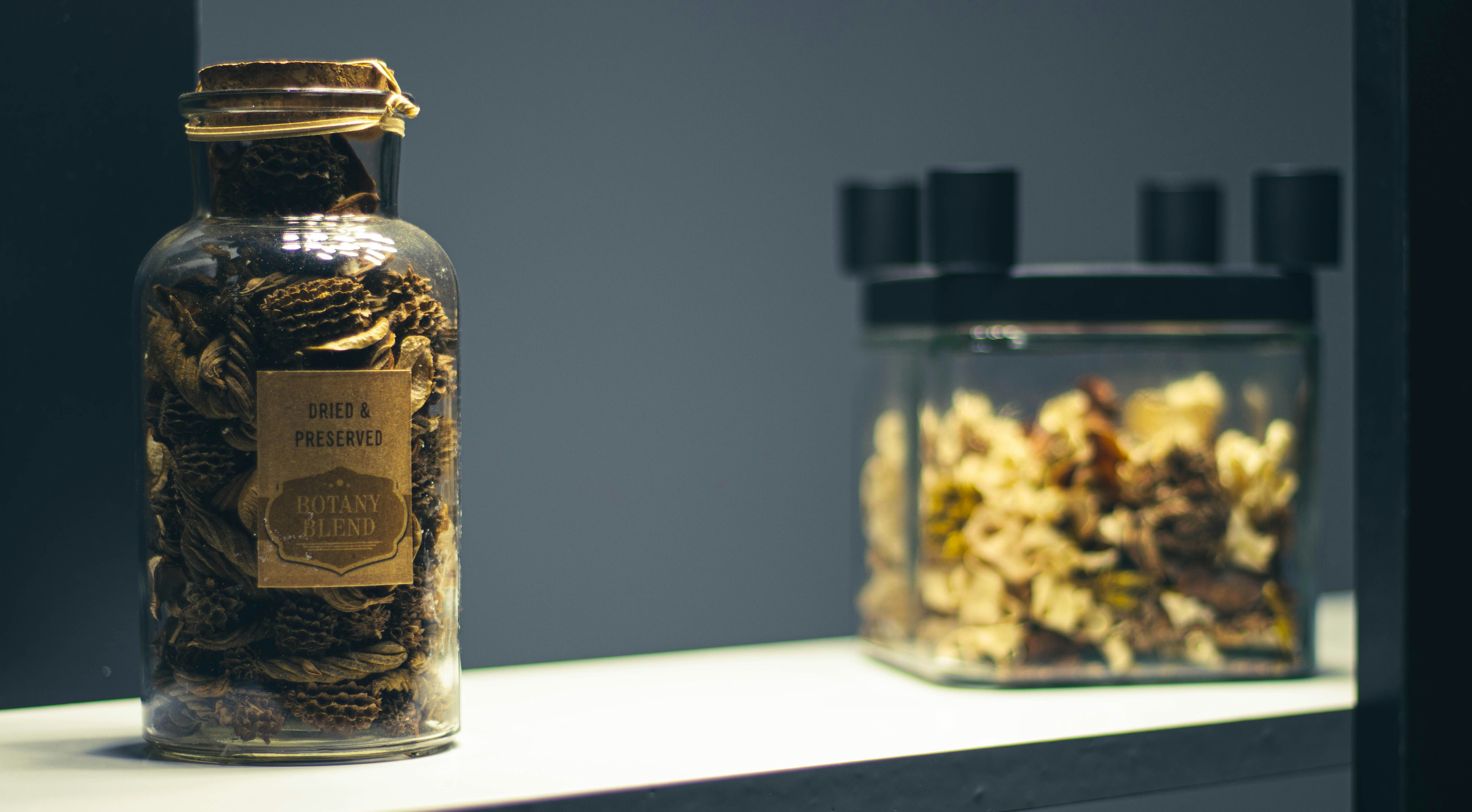 Assorted dried herbs and botanical ingredients in glass jars arranged on dark wooden shelf with warm candlelight