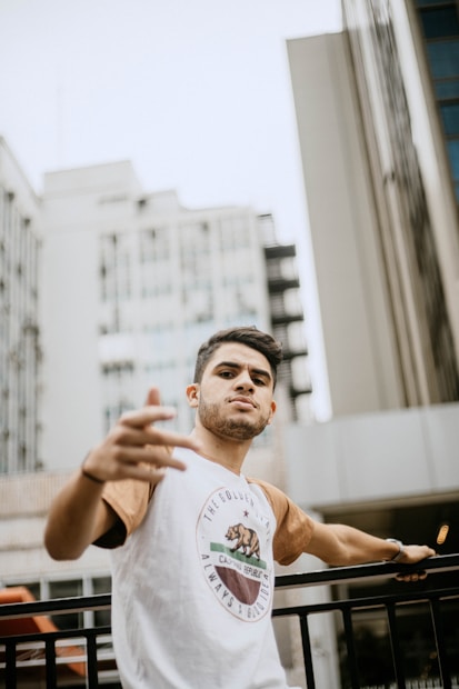 A young man with a confident pose stands in an urban setting. He is wearing a casual t-shirt with a logo, and there's a backdrop of tall buildings. His arm reaches out toward the camera, adding to the dynamic feel of the image.