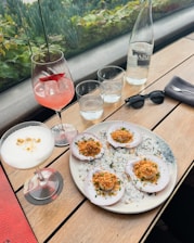 A wooden table set with a gourmet meal featuring scallops served on a plate with a garnish, accompanied by two cocktails in elegant glasses. There is a bottle of water, three empty glasses, a pair of sunglasses, and a fork on a napkin. The background includes a row of lush green plants by a window.