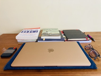 A wooden desk with a neatly arranged layout, including a closed laptop in the center atop a blue sleeve. To the left, a stack of books is positioned, with titles visible. On the right, there is a closed black notebook, a pen with a purple grip, and a pair of glasses. A portable external hard drive is placed to the left of the laptop.