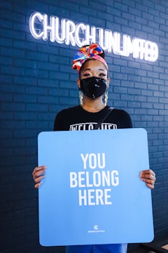 A person wearing a colorful headscarf and black mask holds a blue sign with the words 'YOU BELONG HERE'. The individual stands against a dark brick wall with the illuminated words 'CHURCH UNLIMITED' visible in the background.