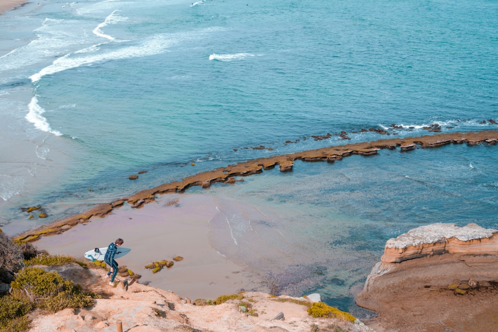 Un hombre sosteniendo una tabla de surf en la cima de una playa rocosa