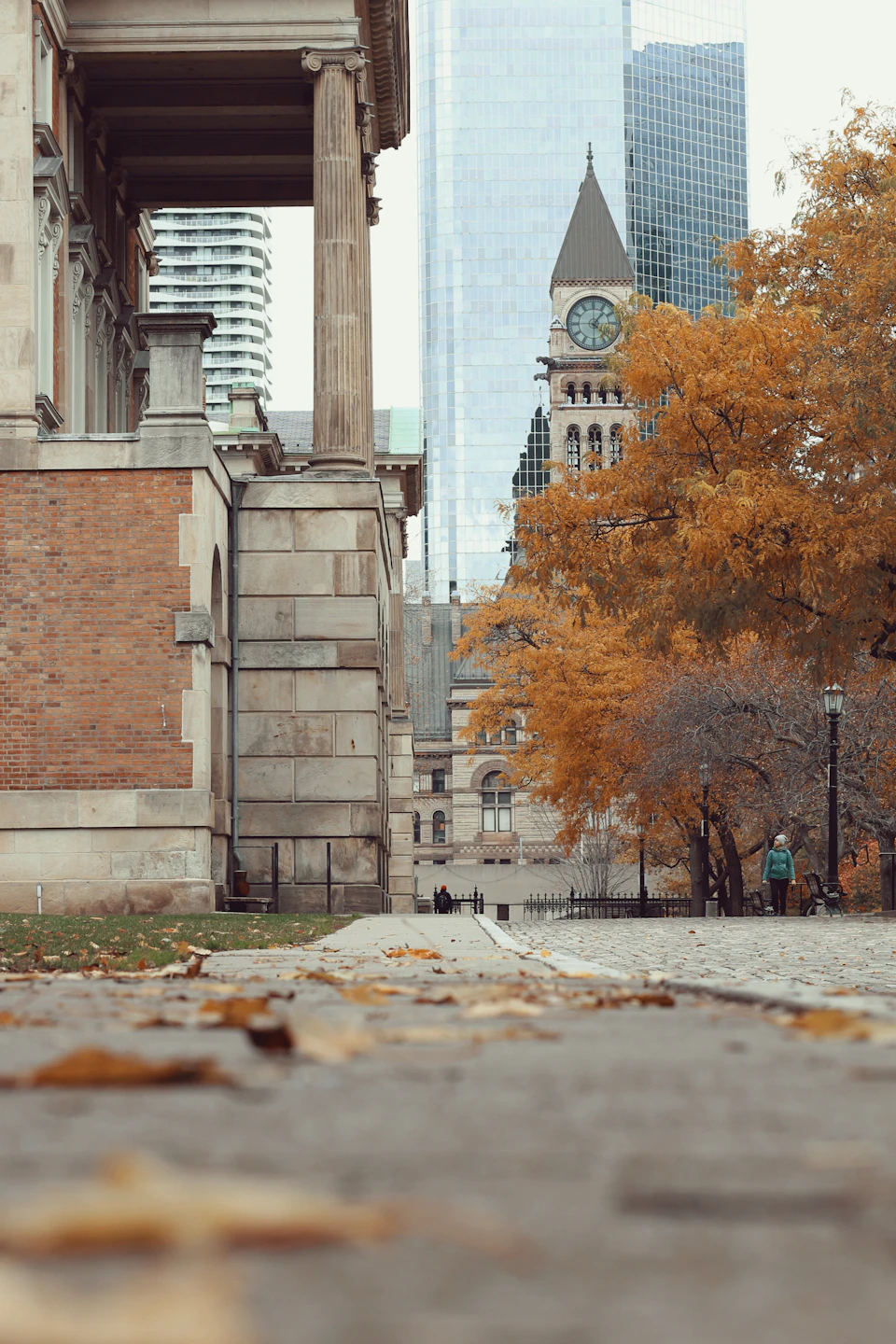 The image depicts a serene autumn scene set against the backdrop of a tall brick building with a clock tower and a smaller clock on its side, situated between two trees adorned with vibrant orange leaves. The perspective is from ground level, looking down a cobblestone street lined with buildings that appear to be in various states of disrepair or abandonment.

There are no discernible texts present in the image.