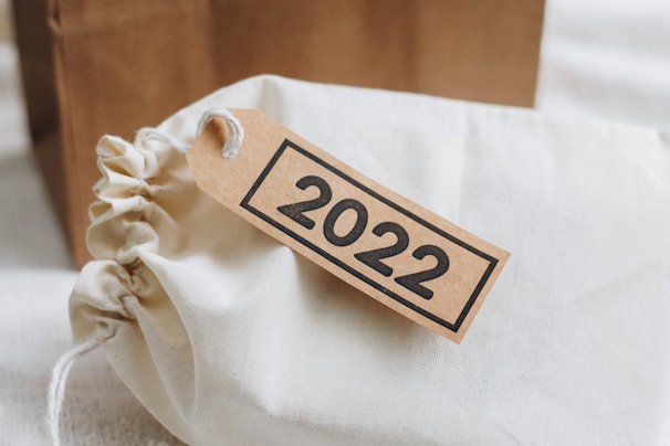 A fabric drawstring bag with a paper tag attached to it displaying the year 2022 in bold black text. The bag is beige or light cream in color, and the tag is brown.