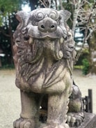 A weathered stone statue of a lion-like mythical creature, often found in East Asian temples. The statue has intricate details on its mane and facial features, displaying a fierce expression. It is situated outdoors, surrounded by a natural setting with trees and a gravel ground.