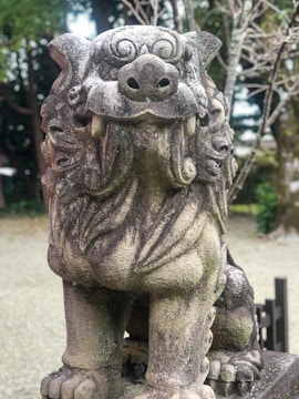 A weathered stone statue of a lion-like mythical creature, often found in East Asian temples. The statue has intricate details on its mane and facial features, displaying a fierce expression. It is situated outdoors, surrounded by a natural setting with trees and a gravel ground.